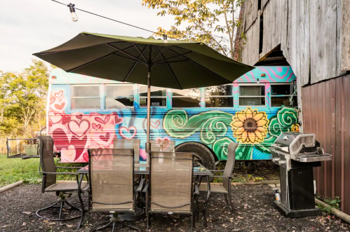 A colorful, painted bus beside a patio table and grill, set in a rustic outdoor space with greenery.