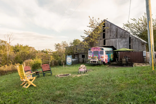 A colorful bus and chairs near a fire pit in front of a rustic barn, surrounded by greenery and trees.