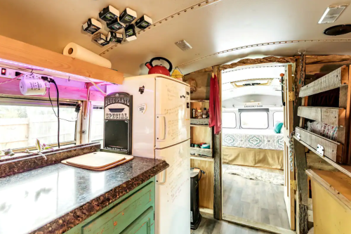Interior of a converted bus featuring a kitchen area with a fridge, countertop, and an open door leading to a cozy bedroom.