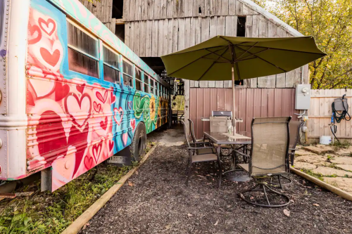 A colorful, graffiti-covered bus parked beside a patio with a table and chairs under an umbrella near a barn.