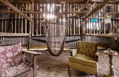 A rustic barn interior featuring a hammock, two vintage chairs, and wooden beams with light streaming through.