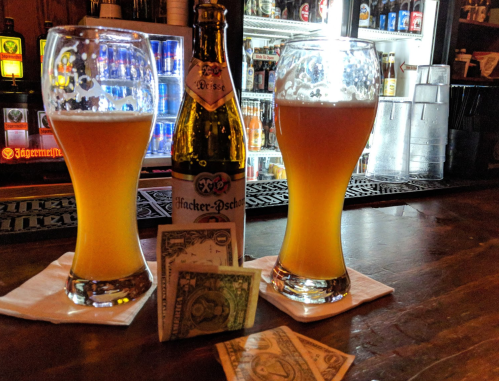 Two glasses of beer beside a bottle, with a few dollar bills on a bar counter. Background shows a variety of drinks.