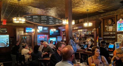 A lively bar scene with patrons enjoying drinks, wooden decor, and a golden ceiling. TVs display sports in the background.