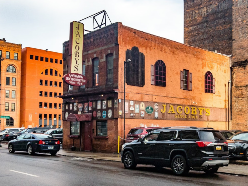Historic building with a sign reading "Jacoby's" and parked cars in front, set against a city backdrop.