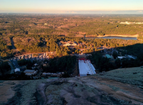 Aerial view of a landscape featuring trees, a lake, and a parking area, with a shadowed foreground.