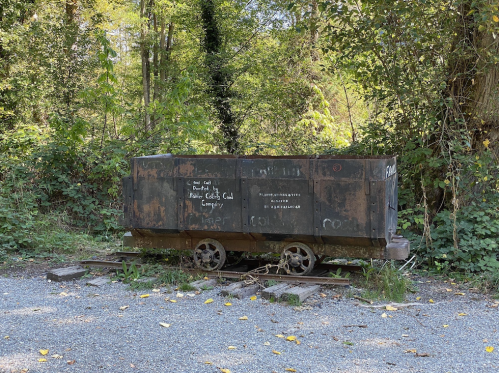 An old, rusty coal cart on tracks, surrounded by trees and gravel, showing signs of wear and age.