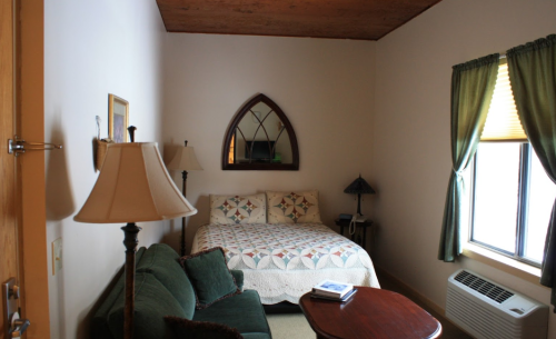 Cozy bedroom with a bed, green curtains, a small table, and lamps, featuring a mirror and an air conditioning unit.