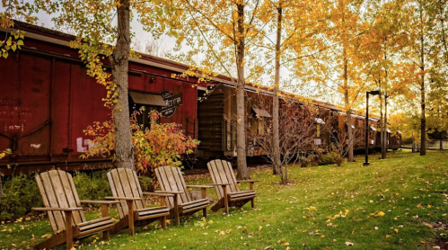 Four wooden chairs sit on a grassy area beside a row of red train cars, surrounded by autumn trees with yellow leaves.