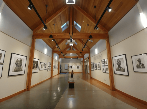 A spacious gallery with wooden beams, featuring black-and-white portraits on the walls and a person sitting in the center.