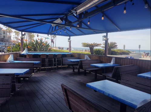 Outdoor dining area with blue tables and a beach view, surrounded by palm trees and a clear sky.
