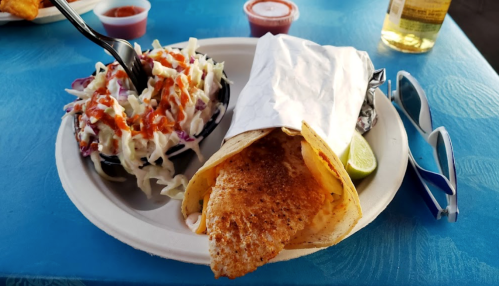 A plate with a fish taco wrapped in paper, coleslaw, lime, and sauces on a blue table. Sunglasses are nearby.