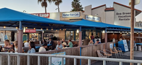 Outdoor dining area of Harbor Fish Cafe with blue awnings, people enjoying meals, and nearby shops in a coastal setting.