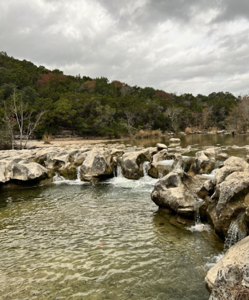 A serene landscape featuring a rocky riverbed with gentle waterfalls, surrounded by trees and cloudy skies.