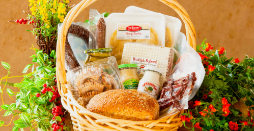 A woven basket filled with various food items, including bread, snacks, and condiments, surrounded by greenery.