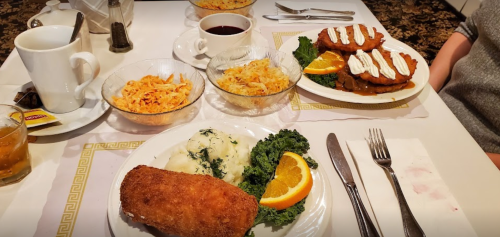 A table set with a hearty breakfast, featuring fried fish, pasta, baked dish, and pancakes, alongside coffee and orange slices.