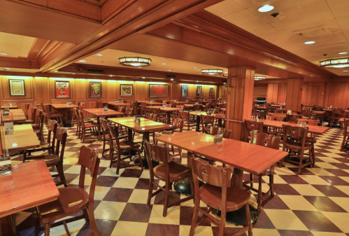 A spacious restaurant interior with wooden tables and chairs, featuring checkered flooring and warm lighting.