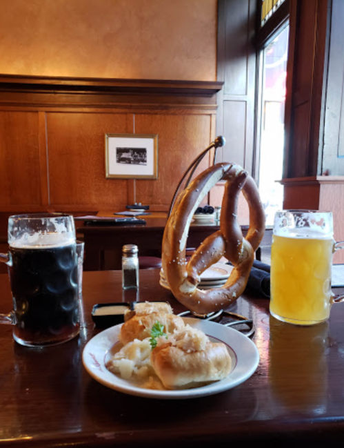 A wooden table with two beers, a large pretzel, and a plate of dumplings topped with sauerkraut.