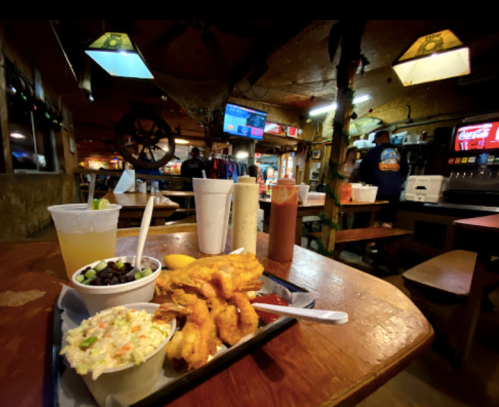 A plate of fried shrimp with coleslaw and black beans, set on a rustic wooden table in a lively restaurant.