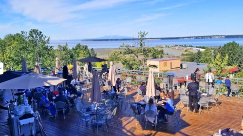 A scenic outdoor dining area with tables, umbrellas, and a view of water and mountains in the background.
