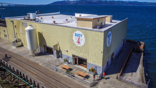 A large yellow building by the water with a sign reading "BUOY," surrounded by a wooden dock and blue water.