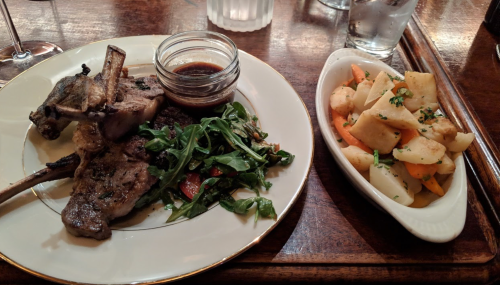 A plate with grilled lamb chops, a side of arugula salad, and a dish of mixed roasted vegetables.