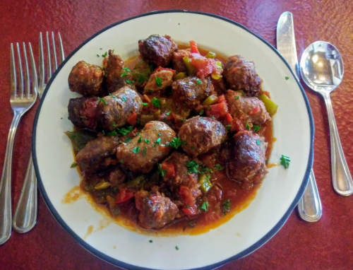 A plate of meatballs in a rich tomato sauce, garnished with herbs, served with a fork and spoon on a red table.