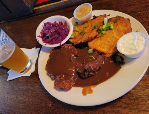 A plate of fried meat with gravy, plantains, purple cabbage, and dipping sauces, alongside a glass of beer.