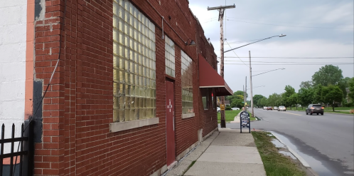 A brick building with glass block windows, an awning, and a sidewalk along a street with cars in the distance.