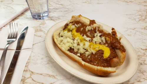 A hot dog in a bun topped with chili, chopped onions, and mustard, served on a plate with utensils nearby.