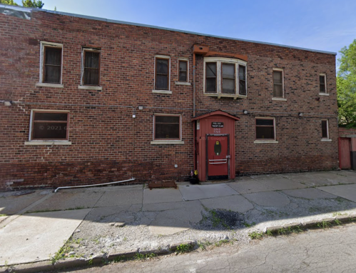 A weathered brick building with a red door and several windows, situated on a cracked sidewalk.