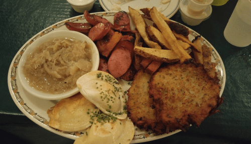 A plate of assorted foods including sausages, fries, sauerkraut, pierogi, and potato pancakes.