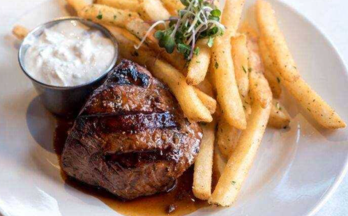 A grilled steak served with crispy fries and a small cup of dipping sauce, garnished with microgreens.