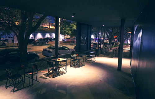 A dimly lit outdoor seating area with empty tables, overlooking a street with parked cars at night.
