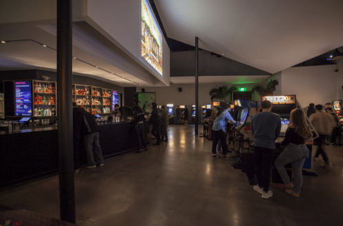 A dimly lit arcade bar with people socializing, vintage arcade machines, and a large screen displaying a game.