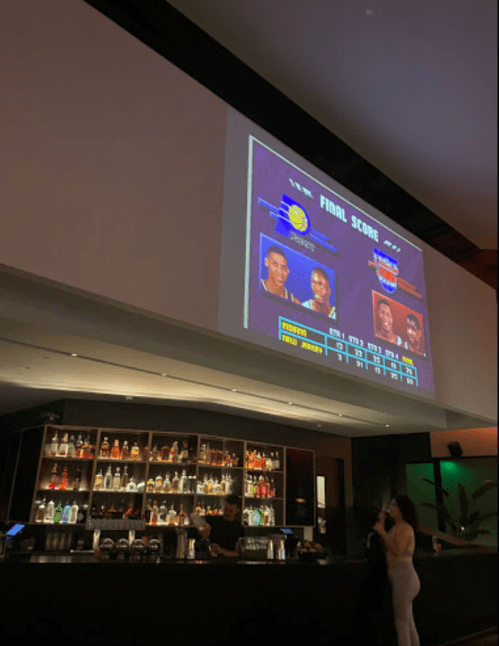 A bar scene with a large screen displaying a basketball game's final score and player stats.