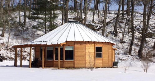 A wooden yurt with a snow-covered roof, surrounded by trees in a winter landscape.