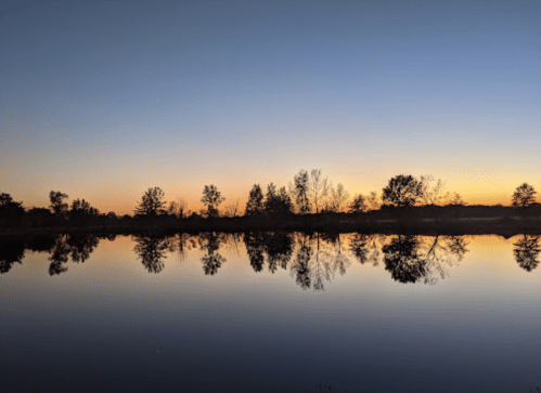 A serene sunset over a calm lake, reflecting trees and a gradient sky from orange to deep blue.