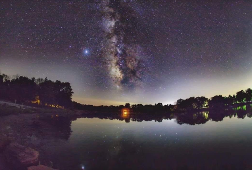 A panoramic view of the night sky filled with stars and the Milky Way, reflecting on a calm lake surrounded by trees.