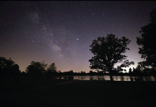 A serene night sky filled with stars and the Milky Way, reflecting over a calm lake, framed by silhouetted trees.