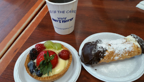 A plate with a fruit tart and a chocolate-dipped cannoli, alongside a coffee cup from Mike's Pastry.