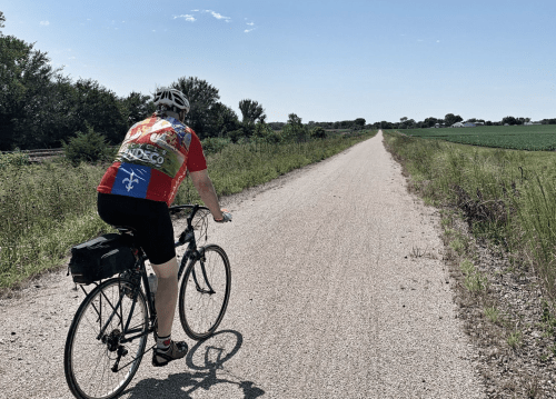 A cyclist in a colorful jersey rides along a gravel path surrounded by greenery and open fields under a clear blue sky.