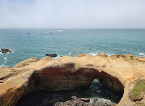 A rocky coastline with a natural arch, overlooking calm blue waters and a foggy sky in the background.