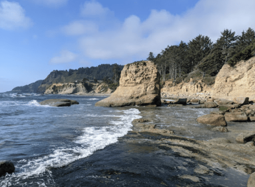 Rocky coastline with waves crashing, surrounded by cliffs and trees under a blue sky with scattered clouds.