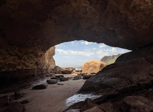 A rocky cave opening reveals a sandy beach and ocean view under a blue sky with scattered clouds.