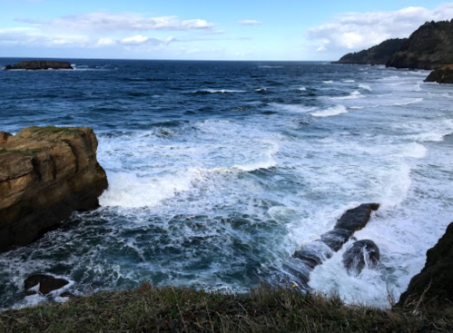 Waves crash against rocky shore under a blue sky with scattered clouds, creating a serene coastal scene.