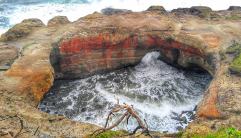 A rocky coastal formation with a circular opening, waves crashing inside, surrounded by rugged terrain and greenery.
