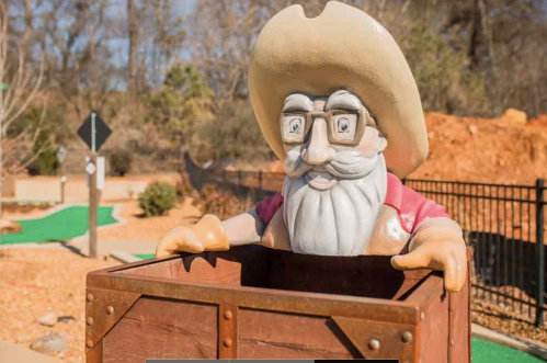 A cartoonish figure of an old man with glasses and a cowboy hat peeks out from a wooden box in a mini-golf setting.