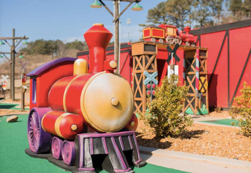 Colorful miniature train structure in a playful outdoor setting, surrounded by greenery and themed buildings.
