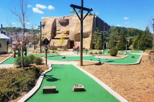 A mini-golf course with green turf, rocky obstacles, and a large stone building in the background under a clear blue sky.