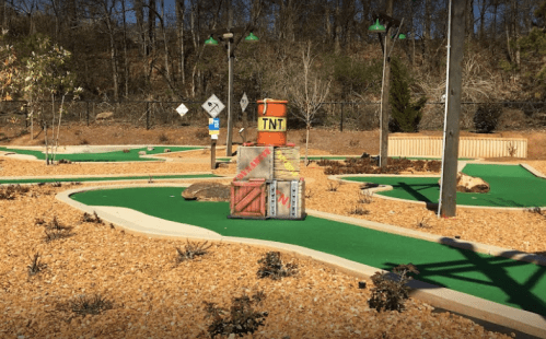 A mini-golf course featuring green turf, gravel paths, and a TNT crate structure surrounded by sparse landscaping.